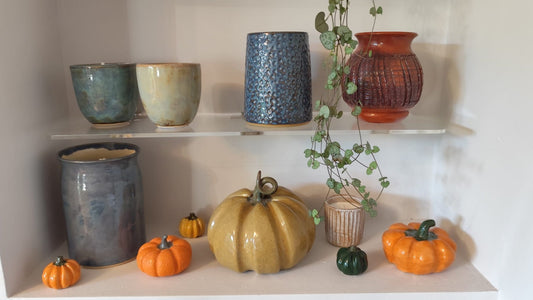 Handcrafted clay mini pumpkins in bright orange and pale yellow glazes arranged on white shelving alongside ceramic pottery and trailing eucalyptus plant, creating an autumn kitchen display